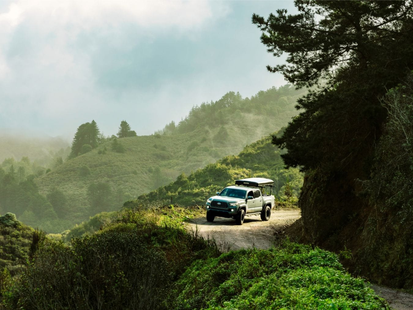 A 3rd gen Tacoma with a rooftop tent on it comes around a dirt road corner.