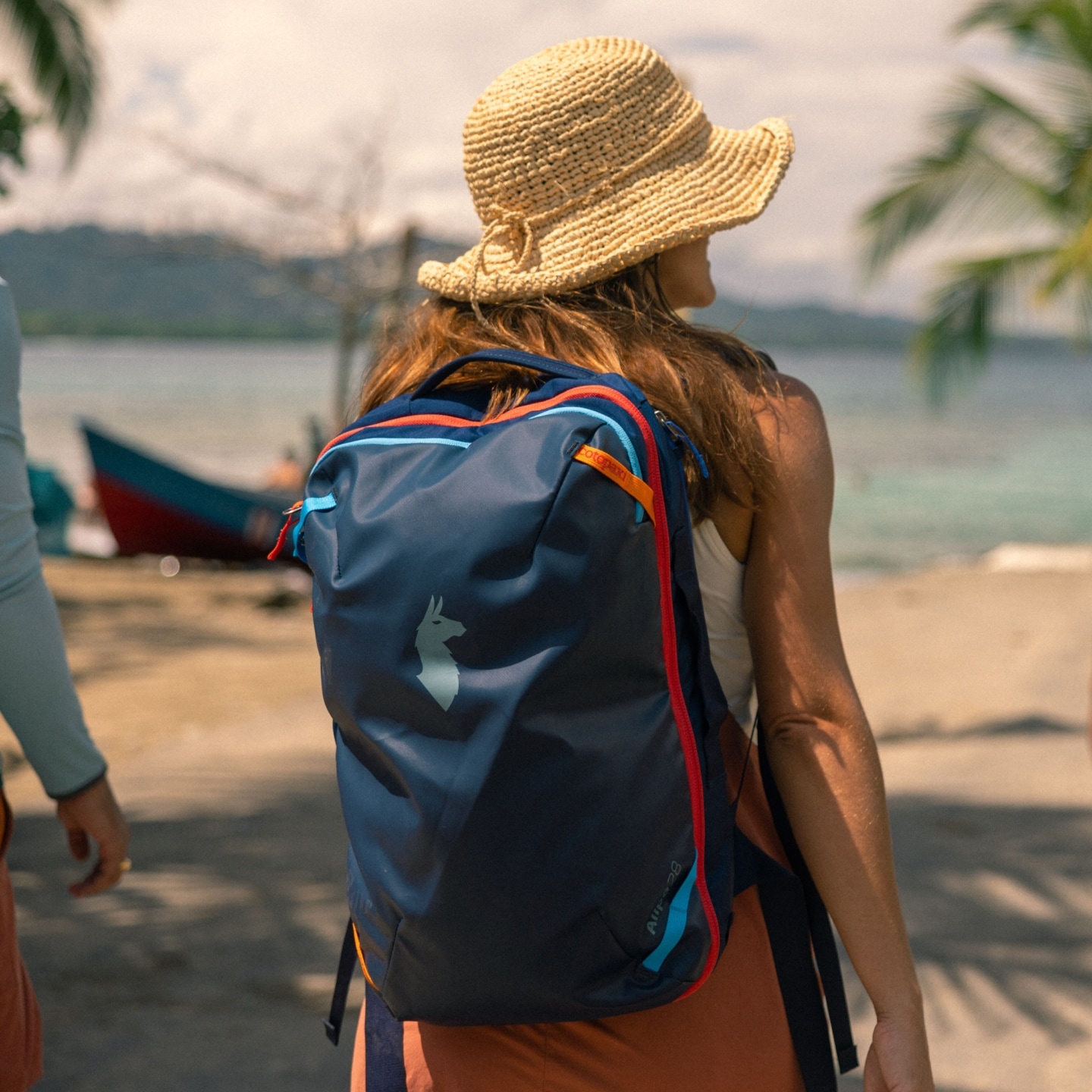 A woman on a beach carrying a blue Cotopaxi backpack.